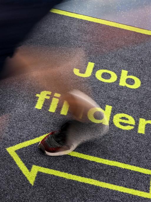 Ein Mann geht in einem Jobcenter über einen Teppich mit der Aufschrift "Job finden". (Gestellte Szene)
