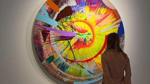 Ein Frau steht in einem Museum vor einem bunten Gemälde von Damien Hirst.
