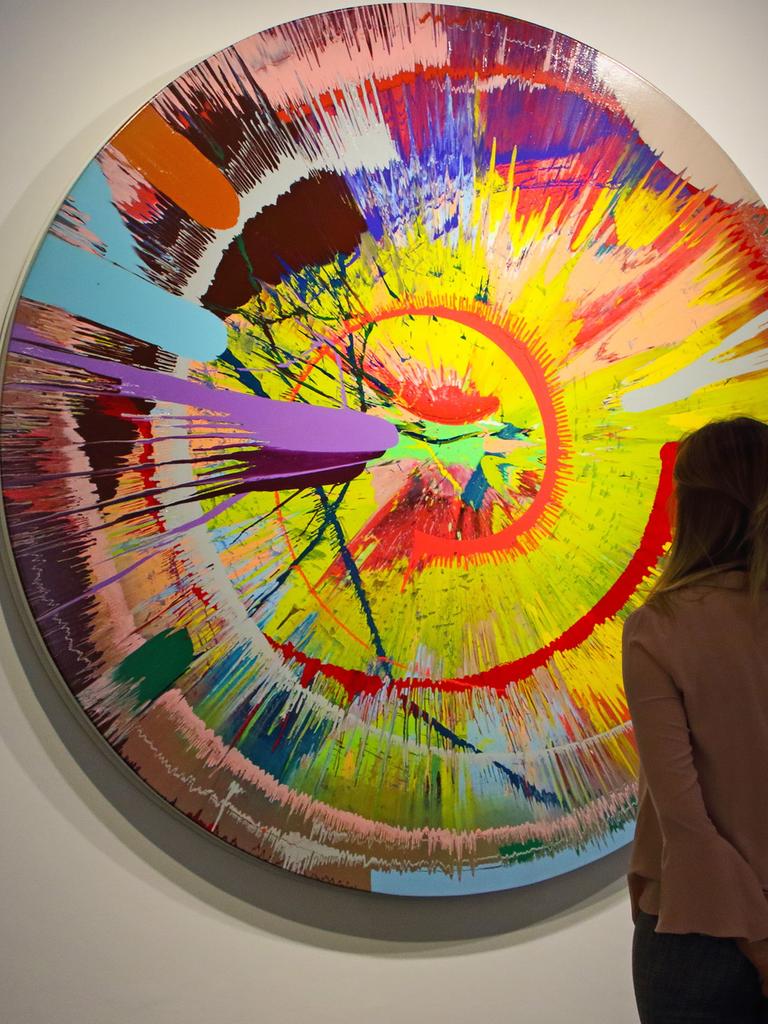 Ein Frau steht in einem Museum vor einem bunten Gemälde von Damien Hirst.