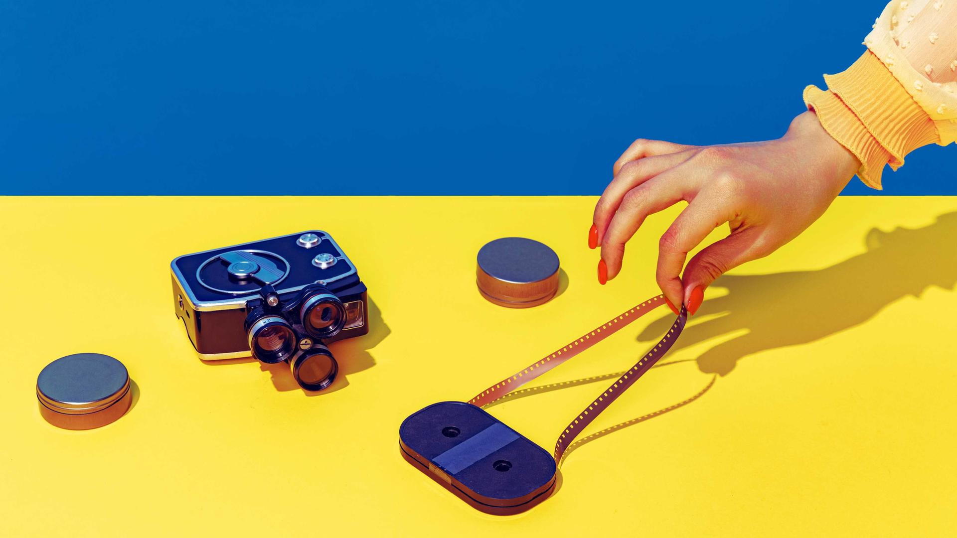 Pop Art Fotografie: Stilisiertes blau-gelbes Bild einer Retro-Kamera und einer Kassette, die eine weibliche Hand mit spitzen Fingern und lackierten Nägeln anfasst.