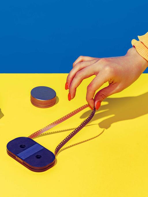 Pop Art Fotografie: Stilisiertes blau-gelbes Bild einer Retro-Kamera und einer Kassette, die eine weibliche Hand mit spitzen Fingern und lackierten Nägeln anfasst.