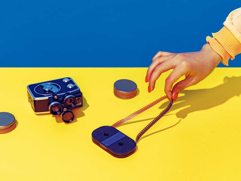 Pop Art Fotografie: Stilisiertes blau-gelbes Bild einer Retro-Kamera und einer Kassette, die eine weibliche Hand mit spitzen Fingern und lackierten Nägeln anfasst.