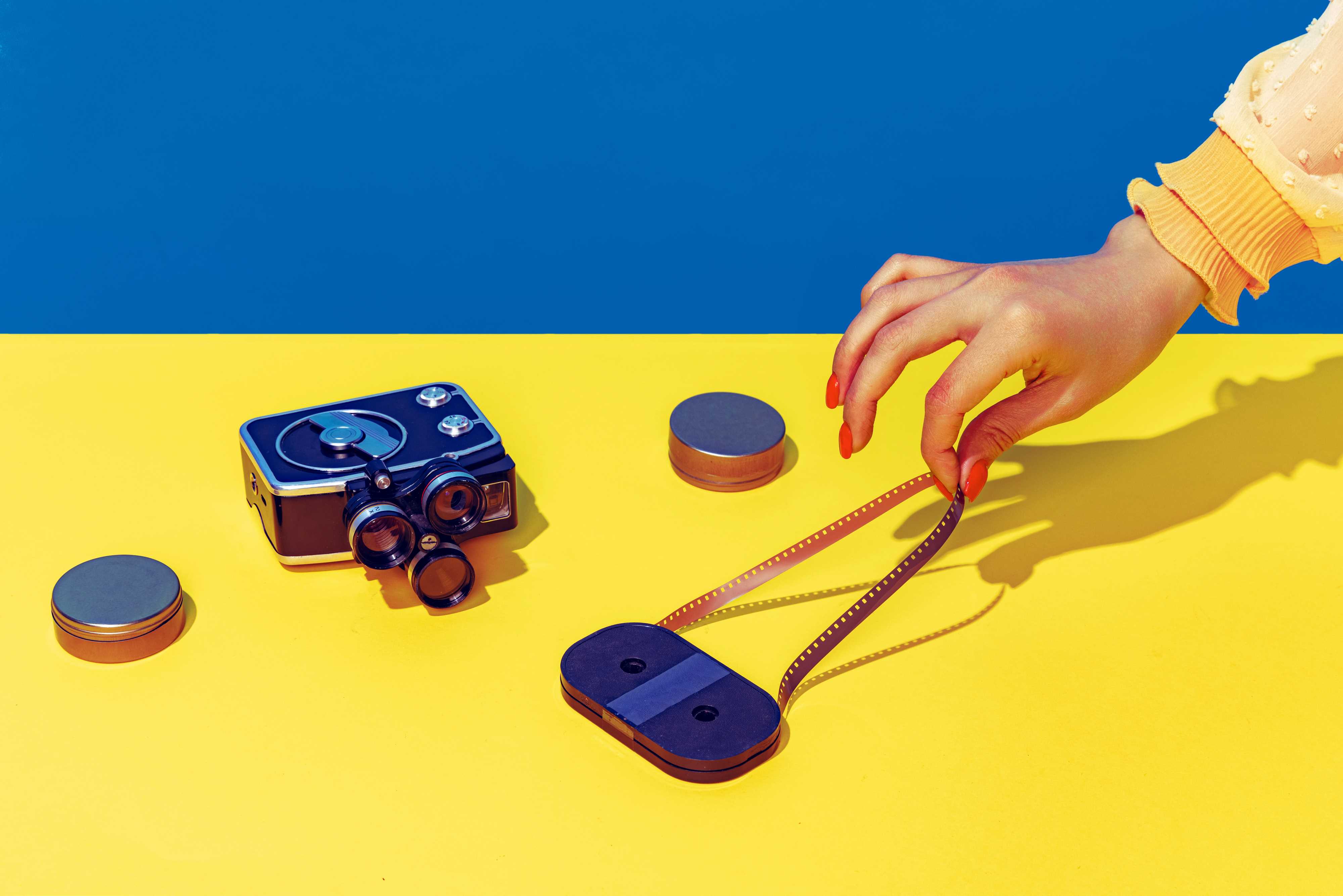 Pop Art Fotografie: Stilisiertes blau-gelbes Bild einer Retro-Kamera und einer Kassette, die eine weibliche Hand mit spitzen Fingern und lackierten Nägeln anfasst.
