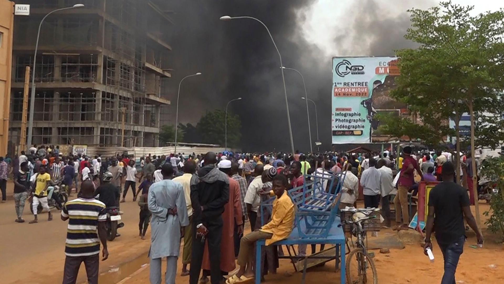 Menschen haben sich auf einem Platz in der Hauptstadt Miamey in Niger versammelt. Im Hintergrund eine große schwarze Rauchwolke.