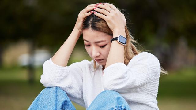 eine Frau sitzt auf einer Wiese. Sie sieht traurig aus und hat die Augen geschlossen. Ihre Hände hat sie auf ihren Kopf gelegt.