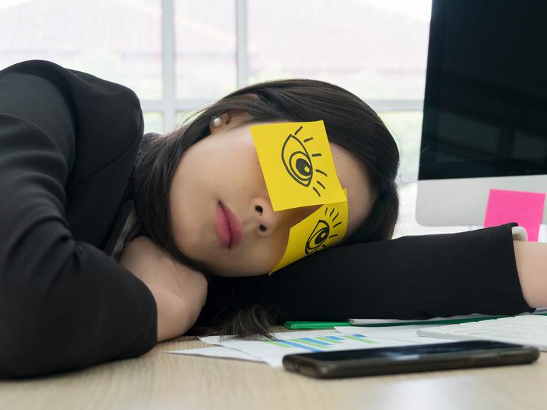 Der Kopf einer jungen Frau mit Zetteln mit aufgemalten offenen Augen im Gesicht liegt auf einem Schreibtisch.