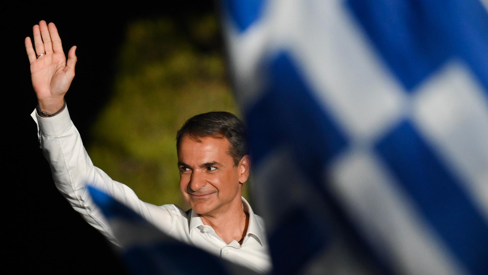 Kyriakos Mitsotakis, Ministerpräsident von Griechenland, winkt während einer Wahlkampfkundgebung vor der Akropolis. Nebendran eine verschwommene Griechenland-Flagge.