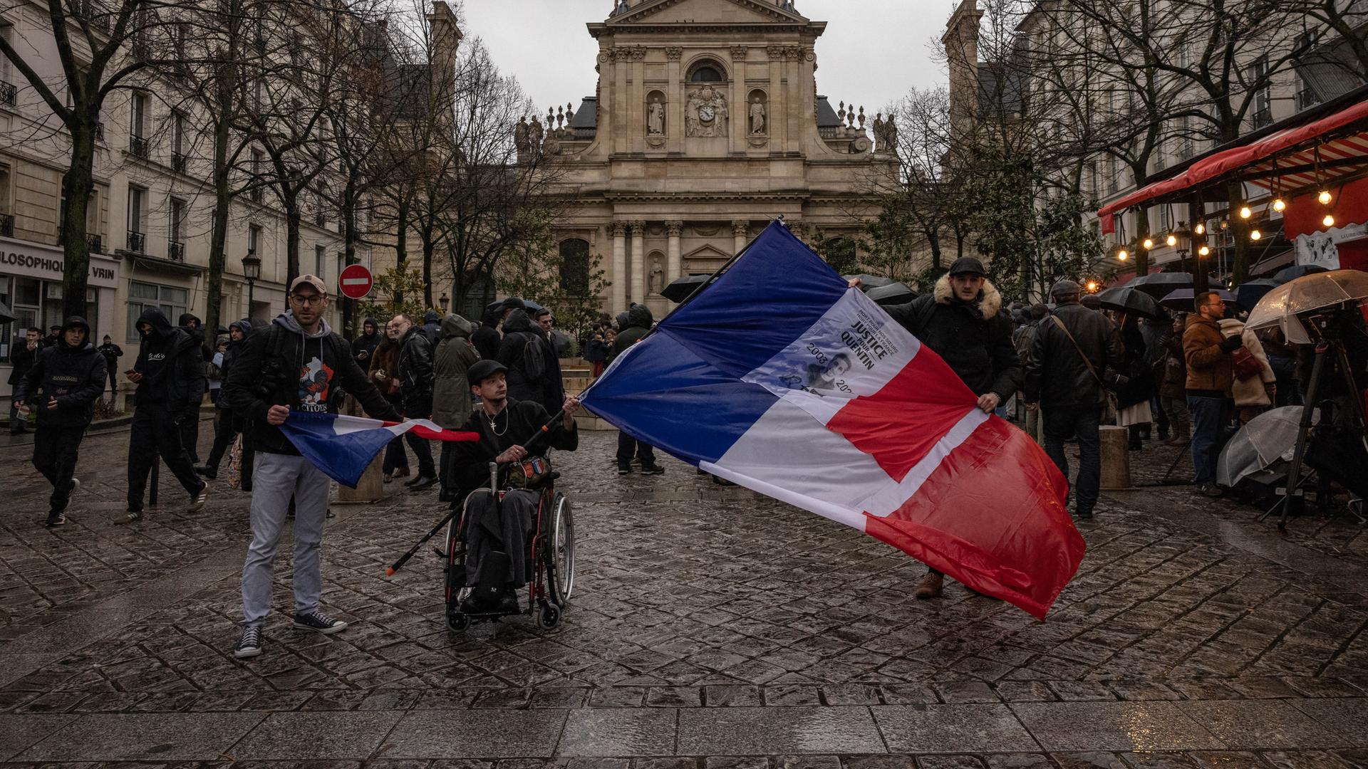 Demonstranten in Lyon halten Fahnen hoch, ganz im Vordergrund ein Rollstuhlfahrer mit einer besonders großen Fahne.
