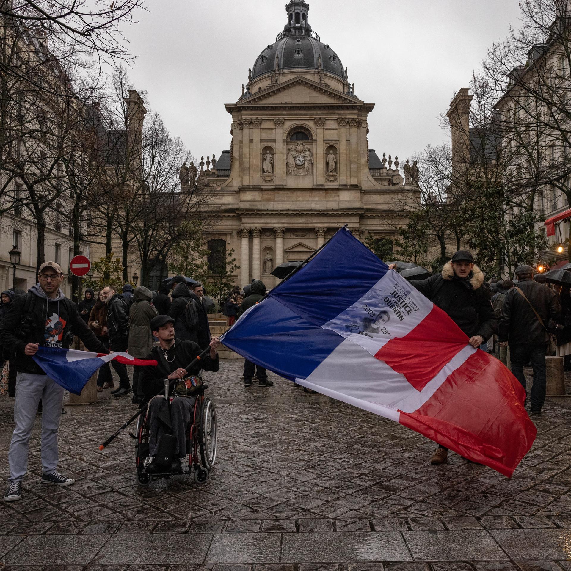 Demonstranten in Lyon halten Fahnen hoch, ganz im Vordergrund ein Rollstuhlfahrer mit einer besonders großen Fahne.