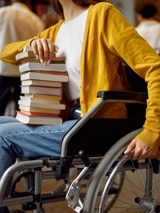 Eine Frau im Rollstuhl hält einen Stapel mit Büchern auf dem Schoß. (Quelle: imago)