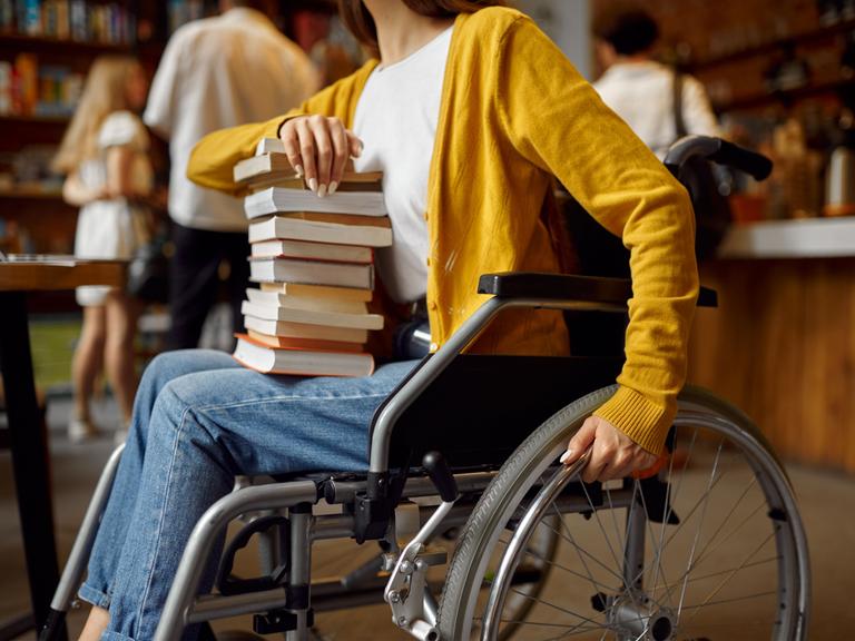Eine Frau im Rollstuhl hält einen Stapel mit Büchern auf dem Schoß. (Quelle: imago)