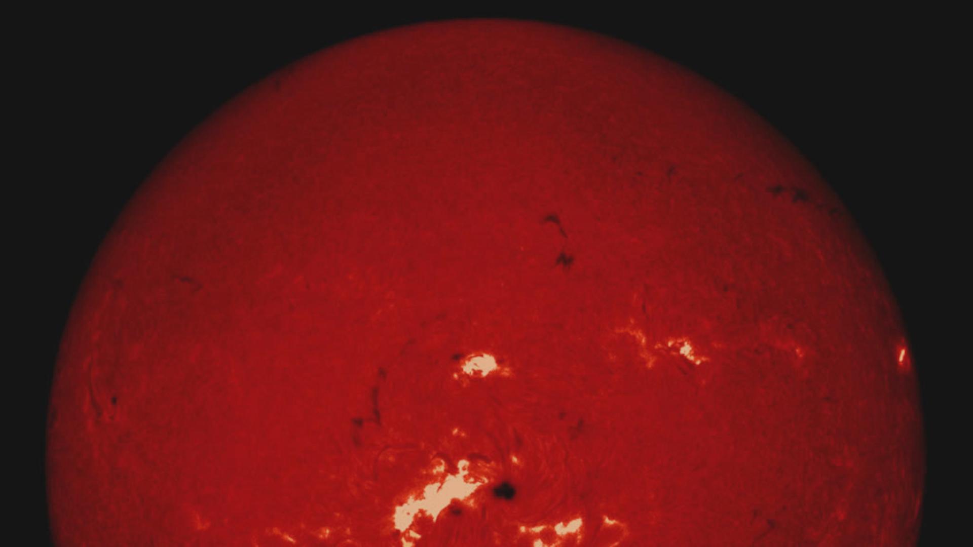Das Bild zeigt eine leuchtend dunkelrote Sonne mit dunklen Flecken und hellen, unregelmäßigen Bereichen. Das Bild zeigt eine leuchtend dunkelrote Sonne mit dunklen Flecken und hellen, unregelmäßigen Bereichen.