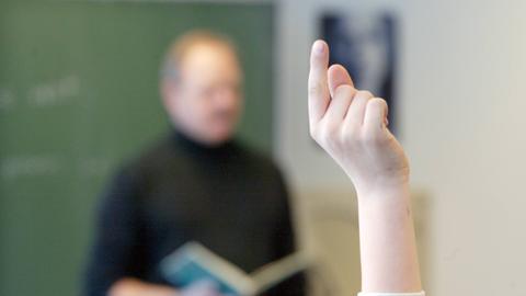 Eine Szene aus einem Klassenraum. Ein Lehrer steht vor einer Tafel und hält ein Buch in den Händen. Vor ihm sind Schüler. Eine Hand, die sich meldet, ist im Vordergrund.
