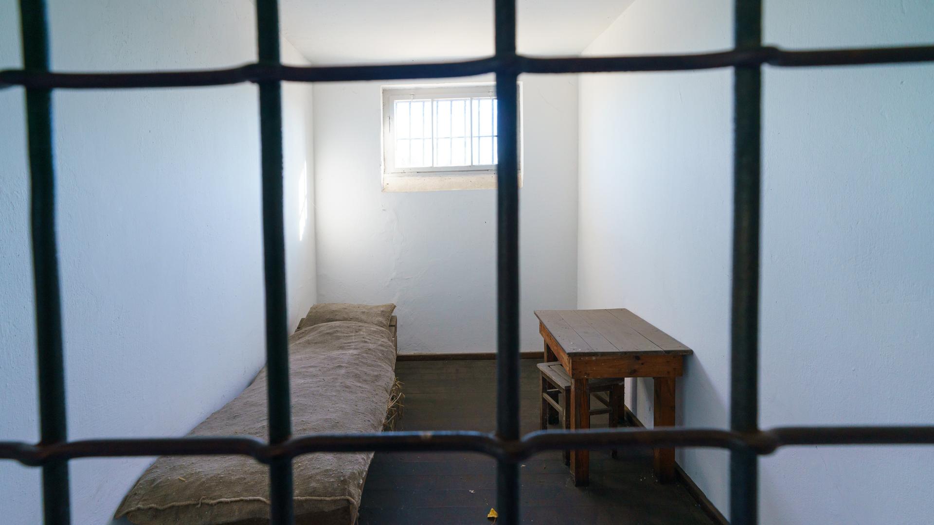 Eine Zelle mit Fenster im Gefängnis des Konzentrationslagers Oranienburg: Hinter Eisengittern sieht man eine Pritsche und einen Holztisch.
