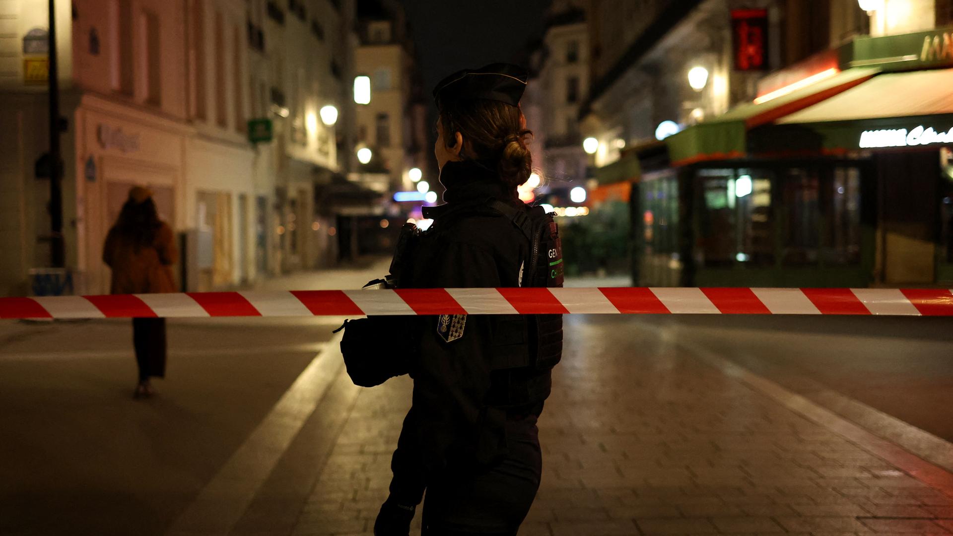 Es ist dunkel. Man sieht die Polizistin als Silhouette. Sie steht in einer Straße hinter einem quer gespannten weiß-roten Flatterband. 
