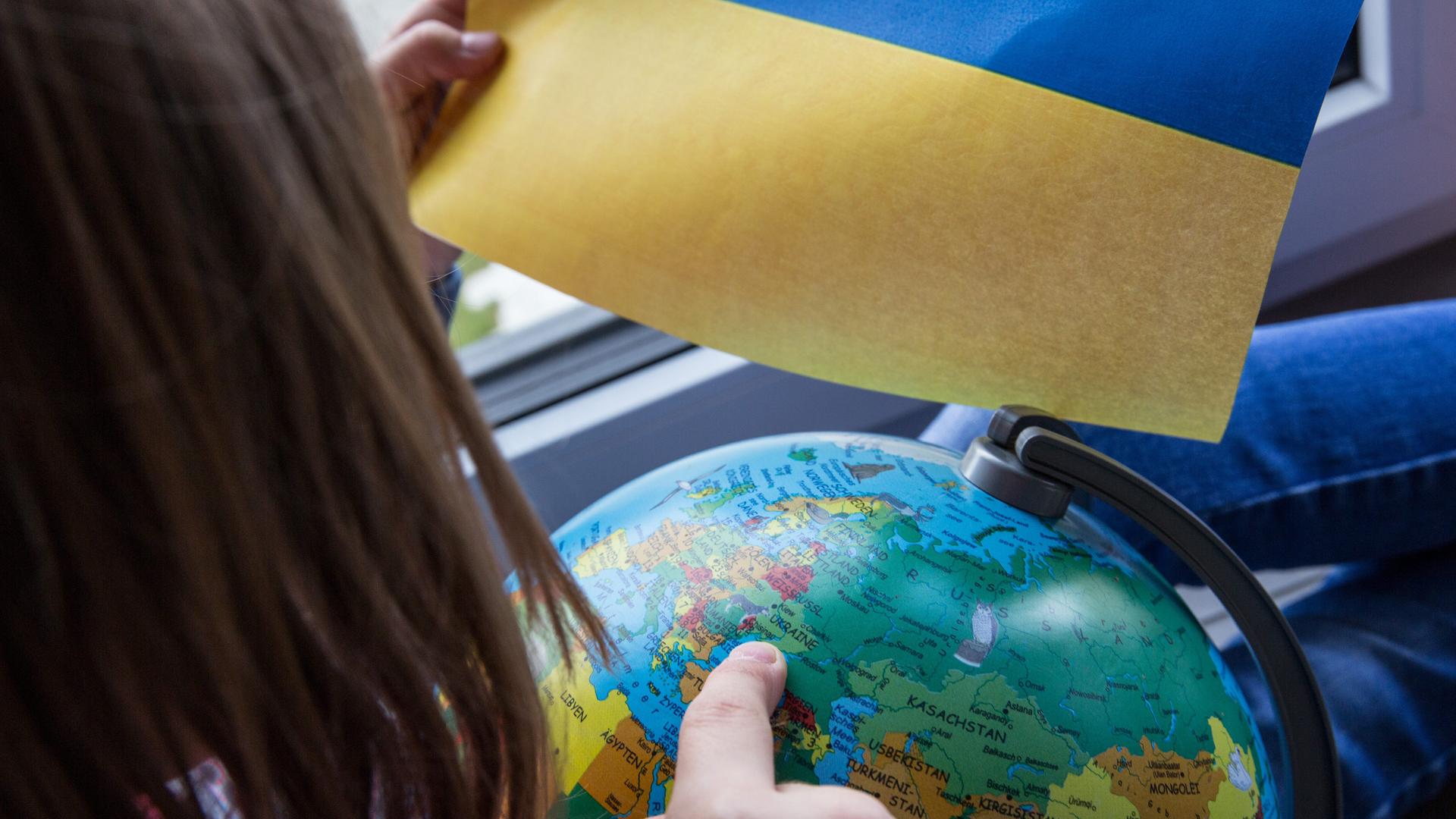 Ein Kind zeigt auf einen Globus und hält eine Ukraine-Flagge in der Hand.