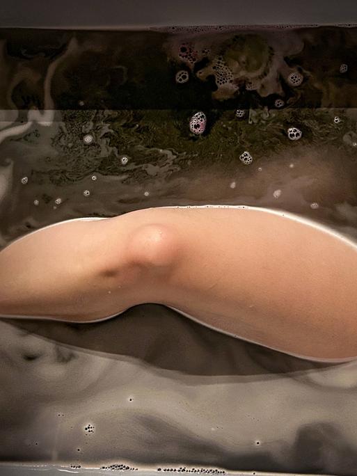 In einer Badewanne ist Wasser eingelassen und darin liegt eine Person, vermutlich eine Frau. Wir sehen ein nacktes Bein, das aus dem Wasser ragt.