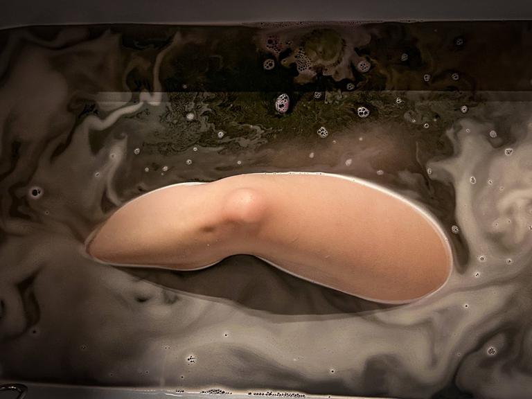 In einer Badewanne ist Wasser eingelassen und darin liegt eine Person, vermutlich eine Frau. Wir sehen ein nacktes Bein, das aus dem Wasser ragt.