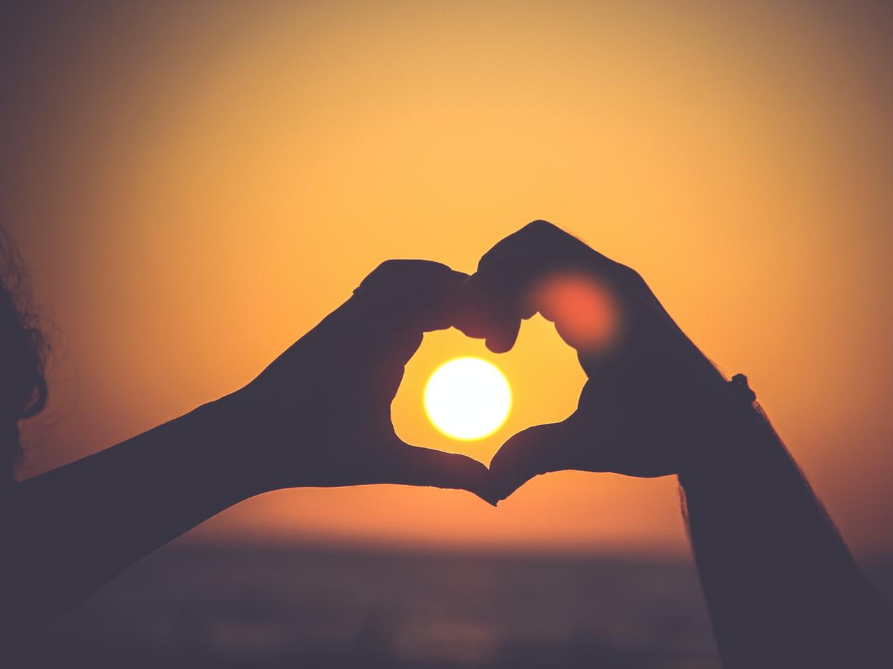 Eine Person bildet mit seinen Händen ein Herz, das die untergehende Sonne umschließt.