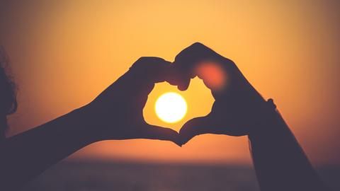 Eine Person bildet mit seinen Händen ein Herz, das die untergehende Sonne umschließt.