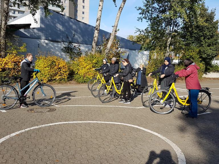 Zu sehen ist ein Schulhof mit weißen Verkehrslinien, vor einer Gruppe Frauen mit Rädern steht eine Trainerin und erklärt.