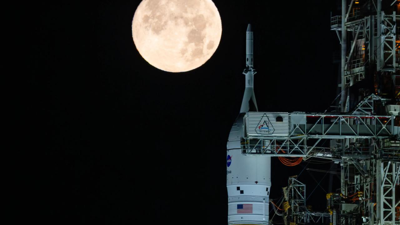 Das Foto zeigt eine weiße Weltraumrakete abends senkrecht auf dem Boden stehend. Im Hintergrund: Ein hell leuchtender großer Vollmond. 