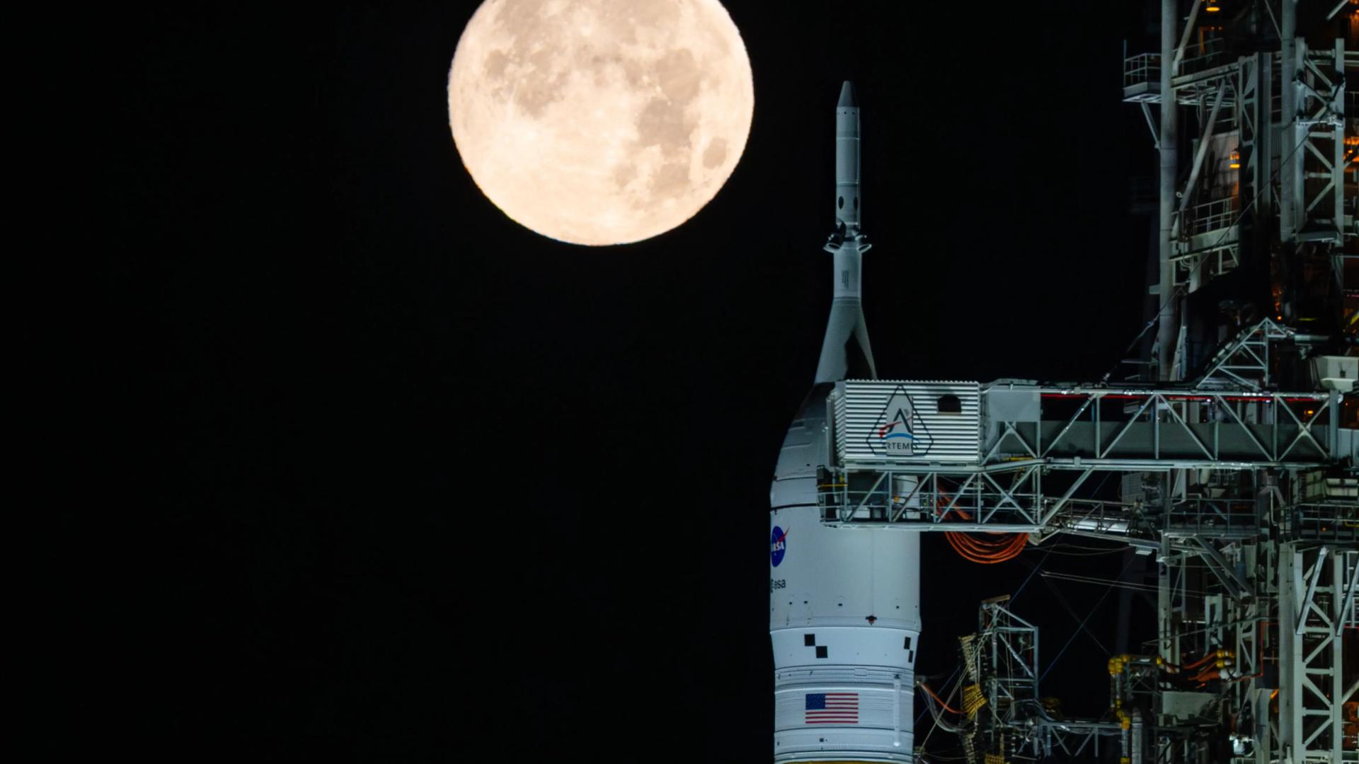 Das Foto zeigt eine weiße Weltraumrakete abends senkrecht auf dem Boden stehend. Im Hintergrund: Ein hell leuchtender großer Vollmond. Das Foto zeigt eine weiße Weltraumrakete abends senkrecht auf dem Boden stehend. Im Hintergrund: Ein hell leuchtender großer Vollmond.