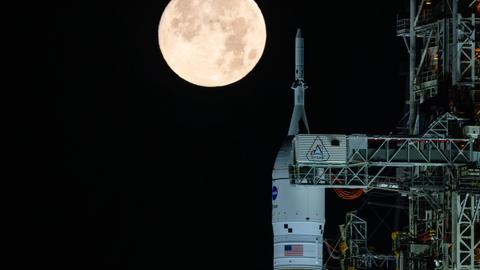 Das Foto zeigt eine weiße Weltraumrakete abends senkrecht auf dem Boden stehend. Im Hintergrund: Ein hell leuchtender großer Vollmond. 