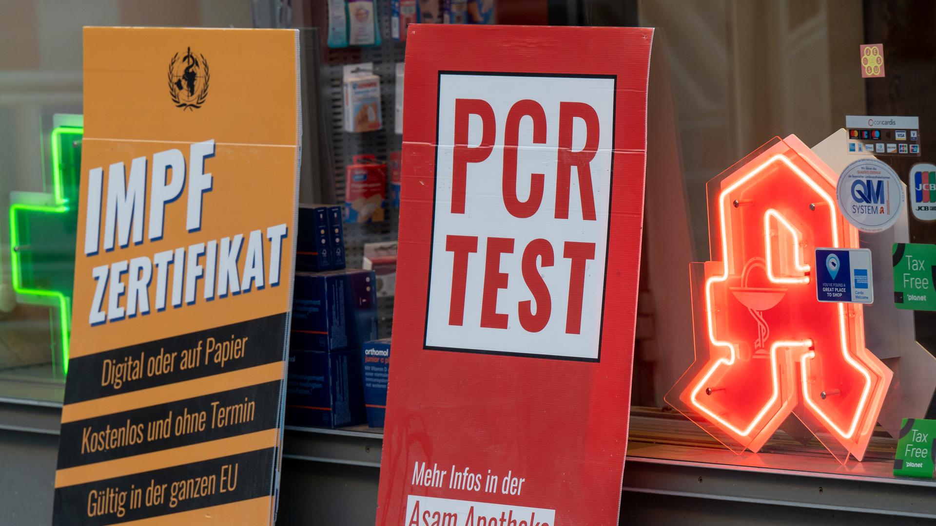 Schilder mit der Aufschrift "Impfzertifikat" und "PCR -Test" stehen vor einer Apotheke in München. 