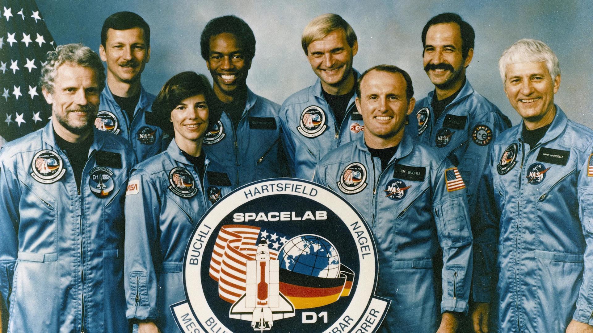 Die Astronauten der D1-Mission stehen in zwei Reihen und lächeln beim Gruppenbild in die Kamera. Vor ihnen steht ein rundes Emblem der Mission mit der Aufschrift "Spacelab" und den Namen der Wissenschaftler.
