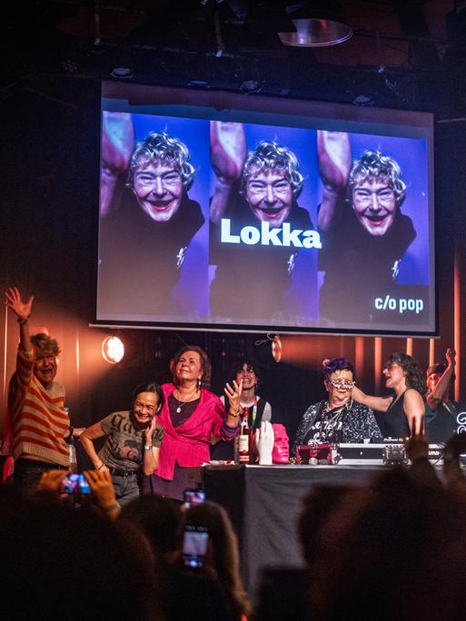 Bei einem Auftritt vor feierndem Publikum stehen mehrere ältere Frauen hinter einem DJ-Pult auf der Bühne.