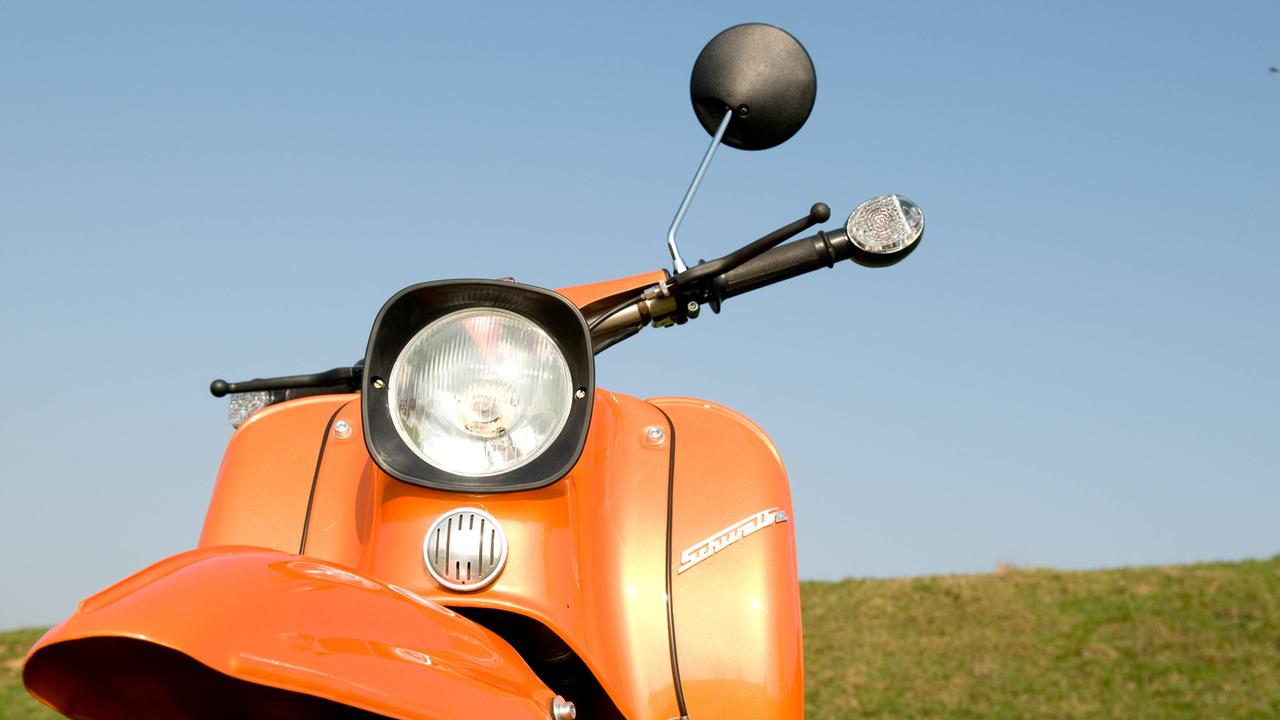 Frontalansicht eines Motorollers der DDR Marke Simson Schwalbe in orange auf einem Feld vor blauem Himmel