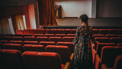 Im Theatersaal: Eine elegante Frau im Kleid steht im Gang zwischen den leeren Zuschauersitzen und blickt nachdenklich gen Theaterbühne.