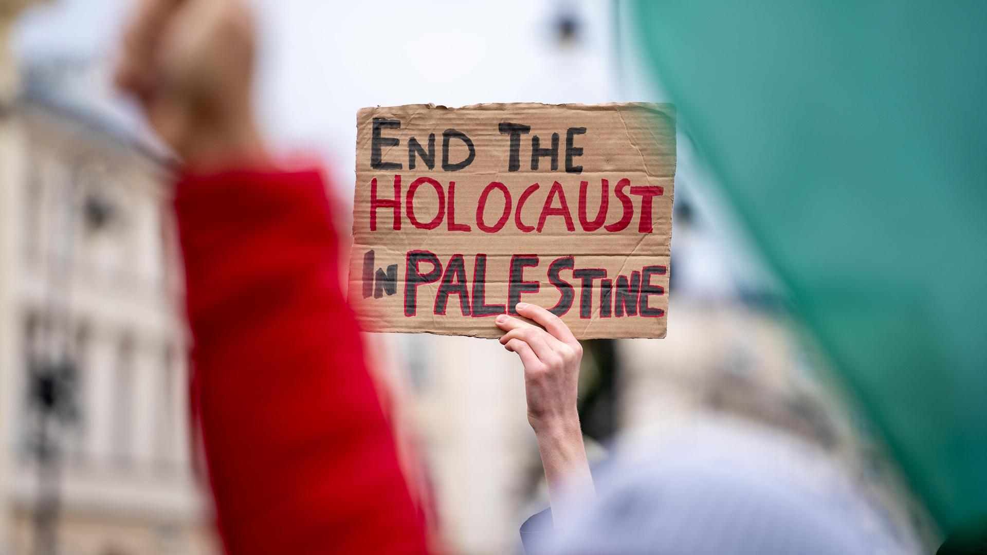 Eine Hand hält ein Schild mit der Aufschrift "Beendet den Holocaust in Palästina" bei einer Demonstration in die Höhe.