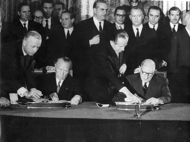 Ein schwarz weißes Foto vom 22. Januar 1963 : Zwei Männer sitzen nebeneinander an einem Tisch und unterzeichnen jeweils ein Dokument. Hinter ihnen stehen sehr viele Männer in Anzügen.