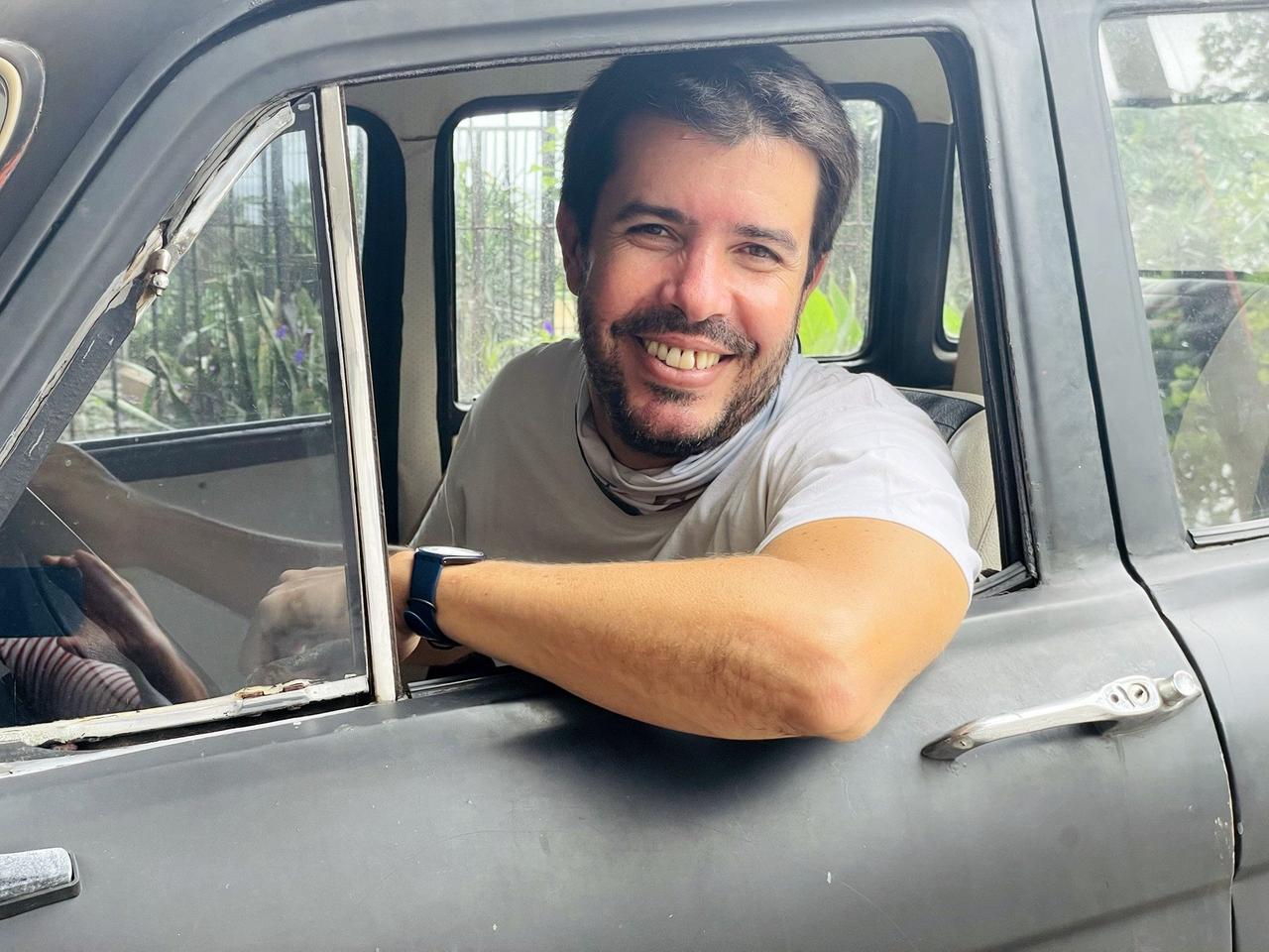Ein junger Mann mit Bart am Steuer eines Autos, den Ellenbogen aus dem Fenster gehängt, lächelt in die Kamera.