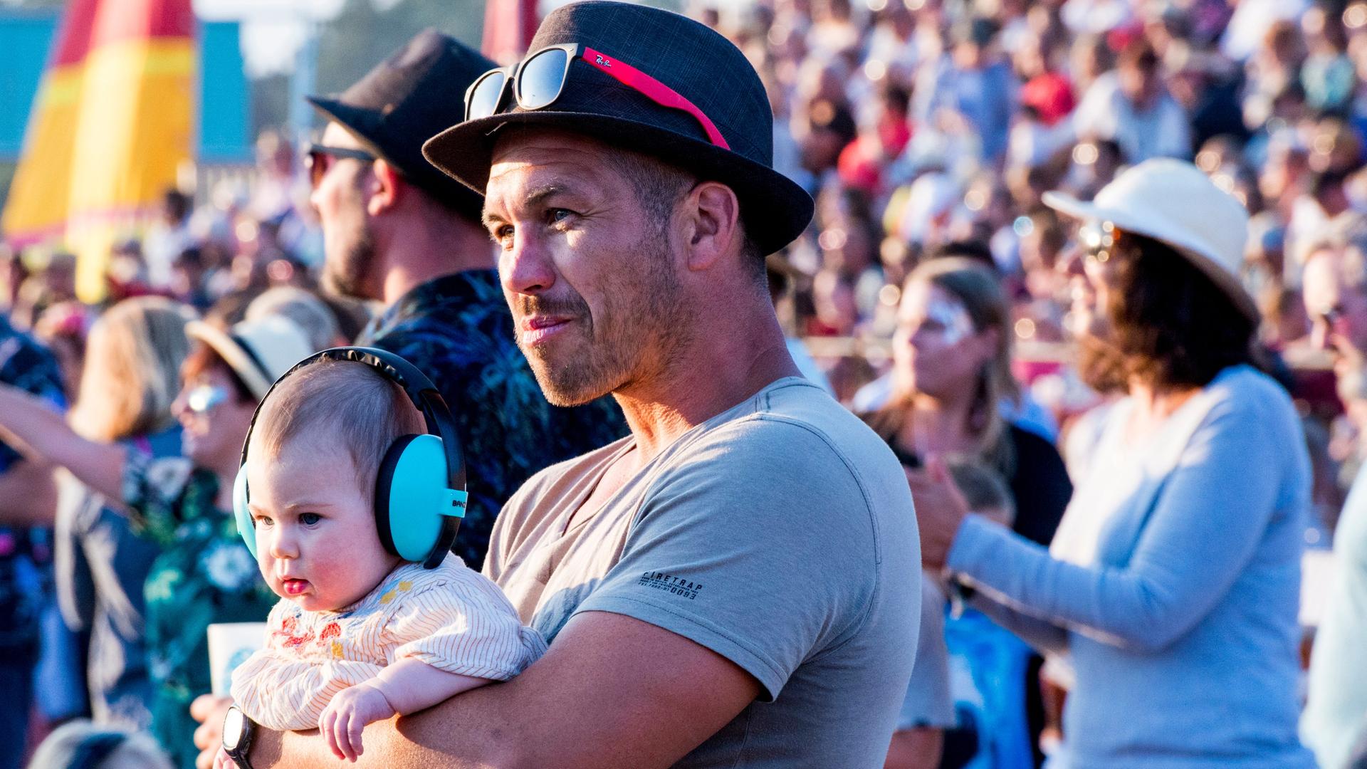 Konzertbesucher beim Latitude-Festival in Suffolk/England: Ein Mann hält ein Baby im Arm, das einen großen Gehörschutz auf den Ohren hat. Konzertbesucher beim Latitude-Festival in Suffolk/England: Ein Mann hält ein Baby im Arm, das einen großen Gehörschutz auf den Ohren hat.