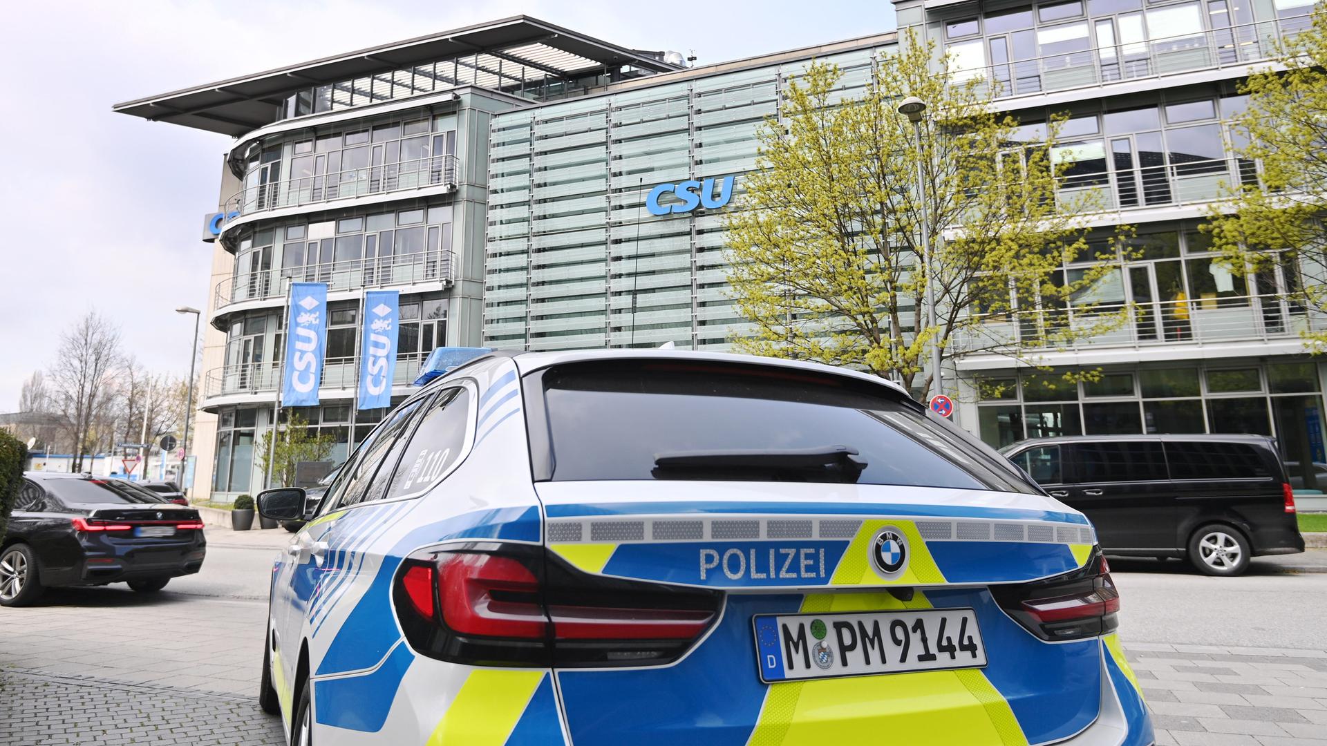 Man sieht das blaue-weiße Polizeiauto in Nahaufnahme von hinten. Dahinter die silbrige große Parteizentrale unter bewölktem Himmel. 
