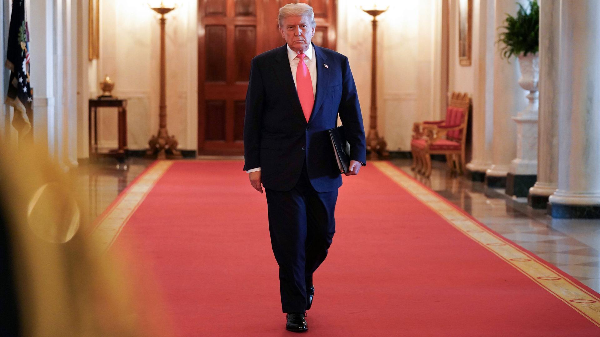 Donald Trump mit Aktenmappe unter dem Arm auf dem roten teppich in einem Korridor des Weißen Hauses