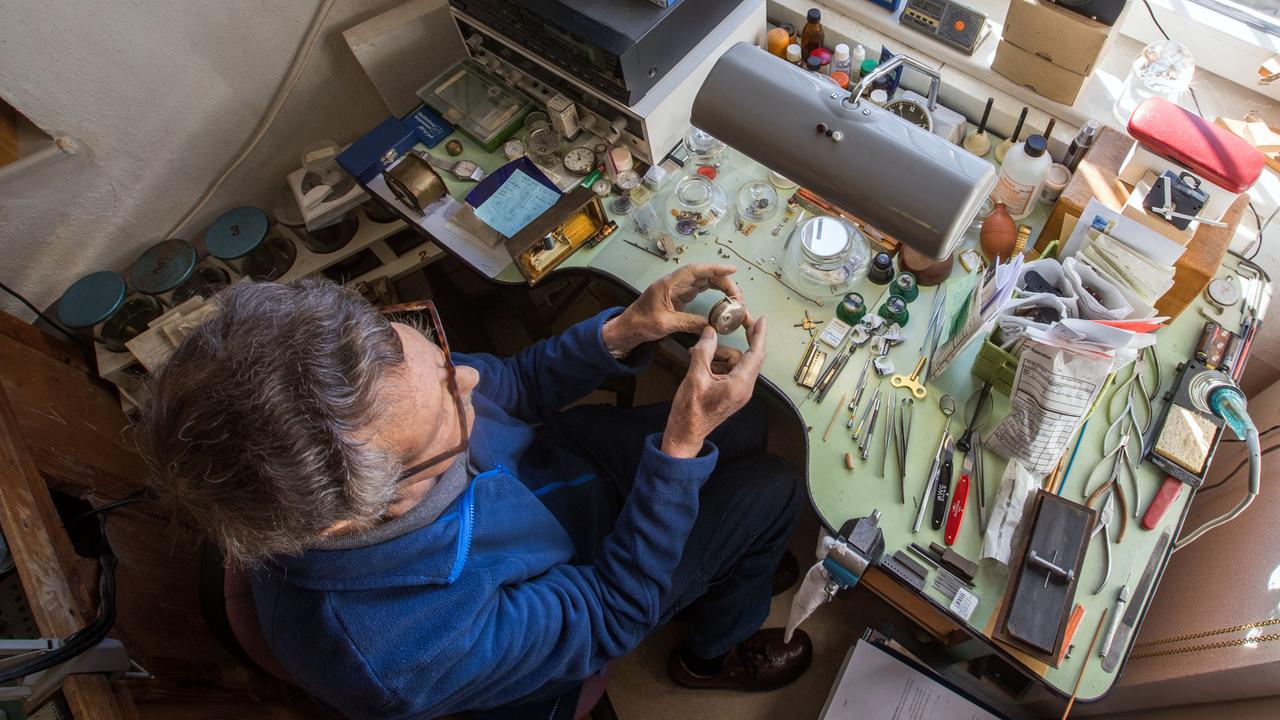 Ein älterer Uhrmachermeister mit ergrautem Haar sitzt in seiner Werkstatt am Arbeitstisch.