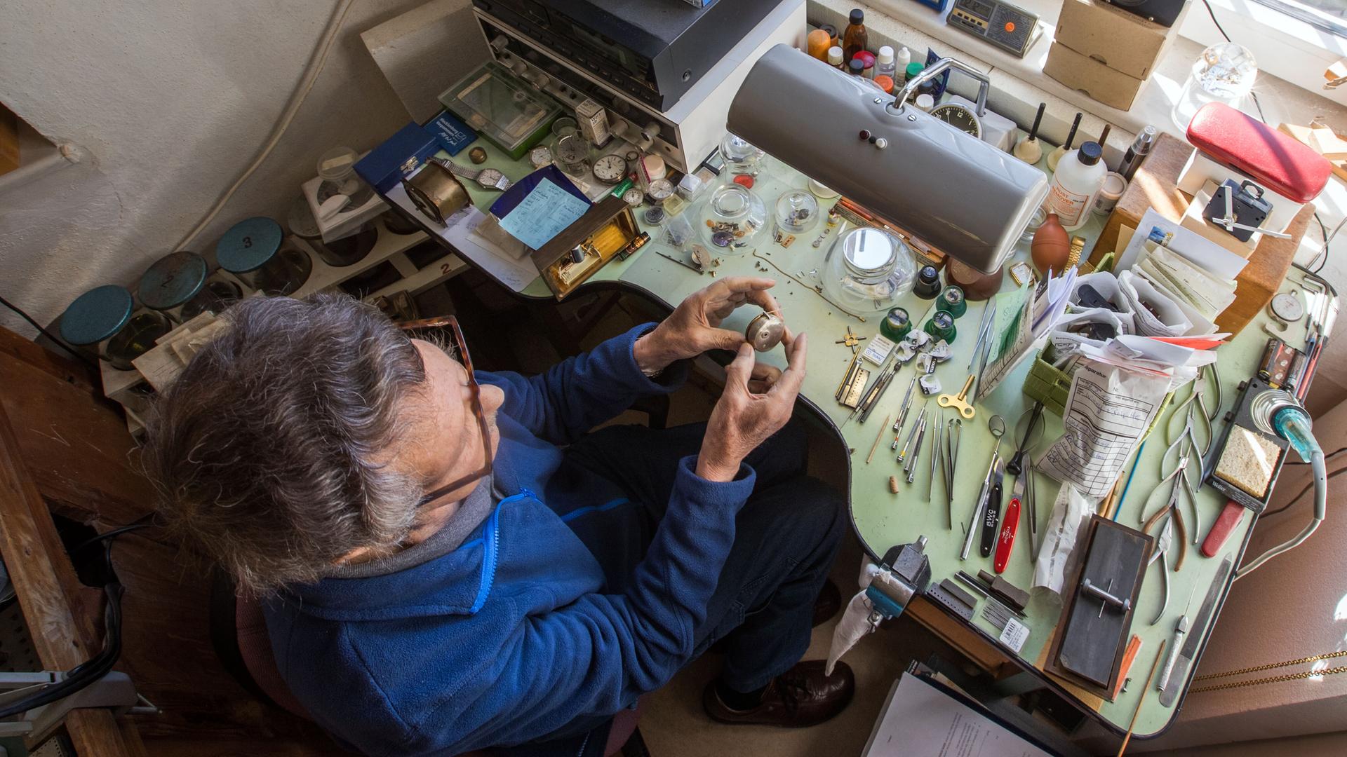 Ein älterer Uhrmachermeister mit ergrautem Haar sitzt in seiner Werkstatt am Arbeitstisch. Ein älterer Uhrmachermeister mit ergrautem Haar sitzt in seiner Werkstatt am Arbeitstisch.