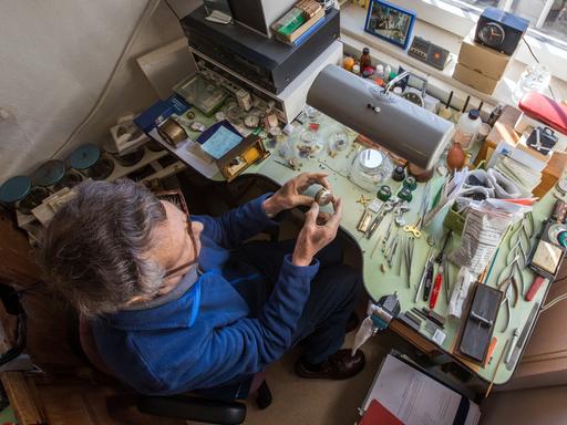 Ein älterer Uhrmachermeister mit ergrautem Haar sitzt in seiner Werkstatt am Arbeitstisch.