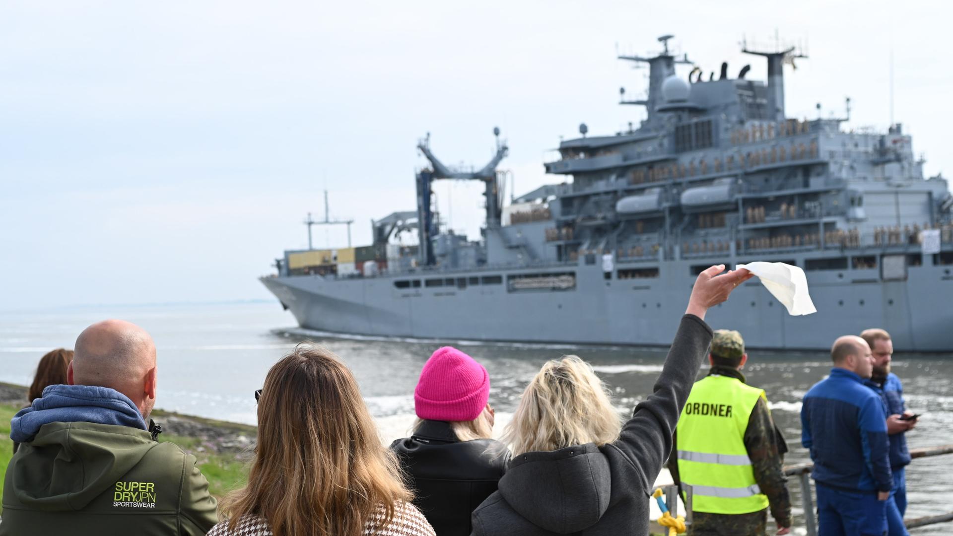 Ein Bundeswehrschiff verlässt einen Hafen, Menschen sehen ihm nach, eine Frau winkt mit einem Taschentuch.