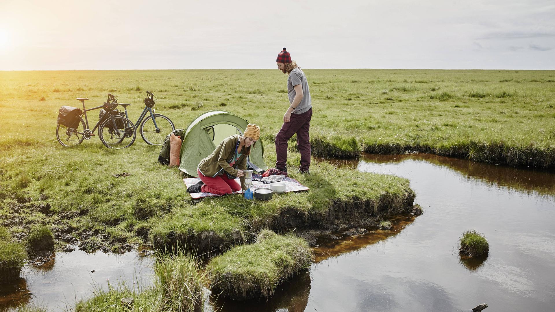 Ein junger Mann und eine junge Frau mit Zelt und zwei Fahrrädern campen auf einer grünen Wiese am Wasser.