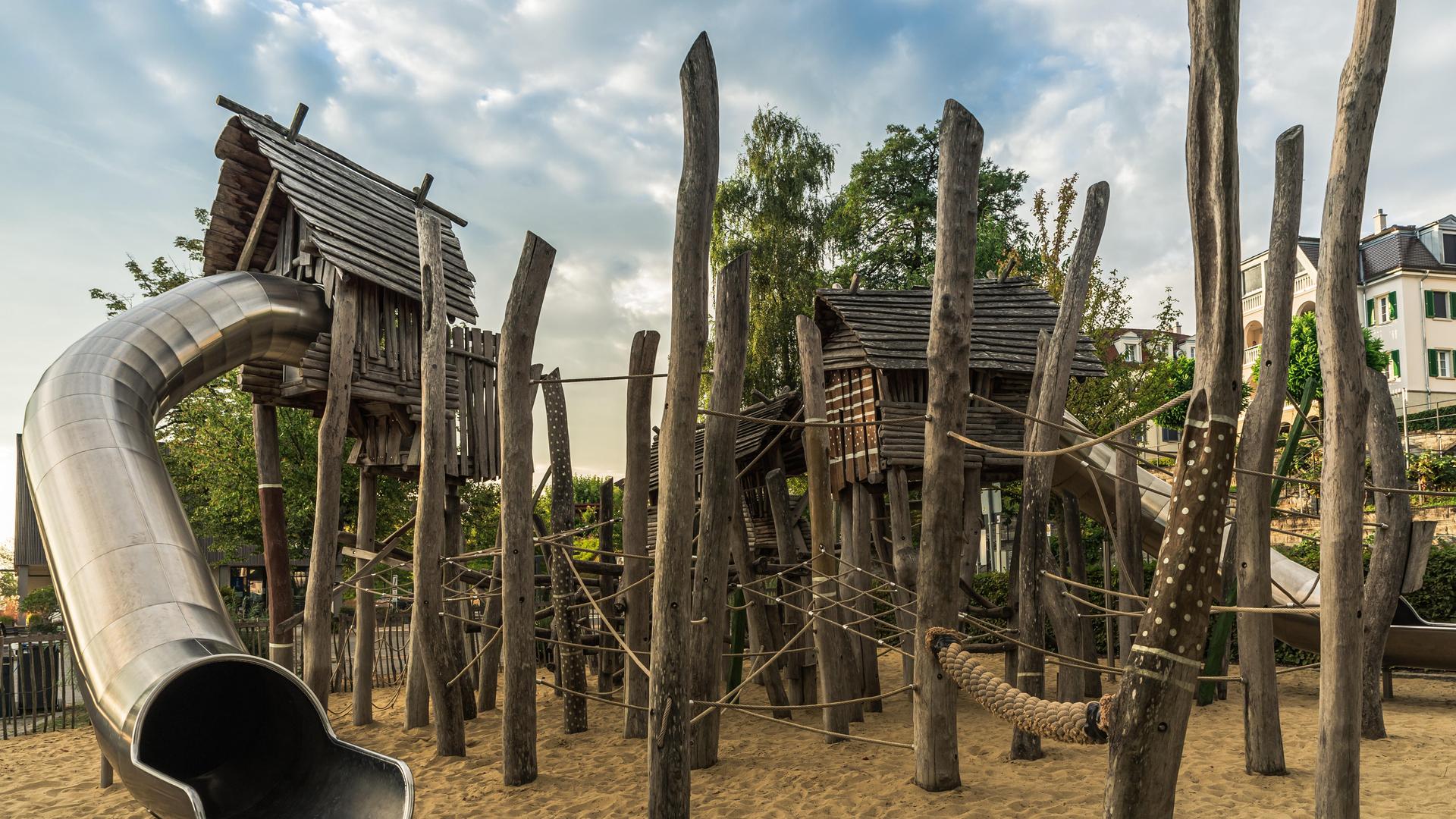 Ein Kinderspielplatz mit Holzhütten, Klettergeräten aus Holzpfählen und großzügiger Rutsche, der an einen Wald erinnert. Ein Kinderspielplatz mit Holzhütten, Klettergeräten aus Holzpfählen und großzügiger Rutsche, der an einen Wald erinnert.