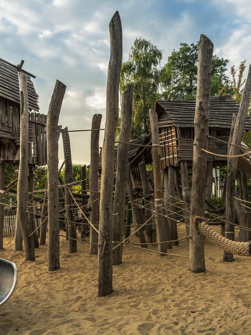 Ein Kinderspielplatz mit Holzhütten, Klettergeräten aus Holzpfählen und großzügiger Rutsche, der an einen Wald erinnert.  