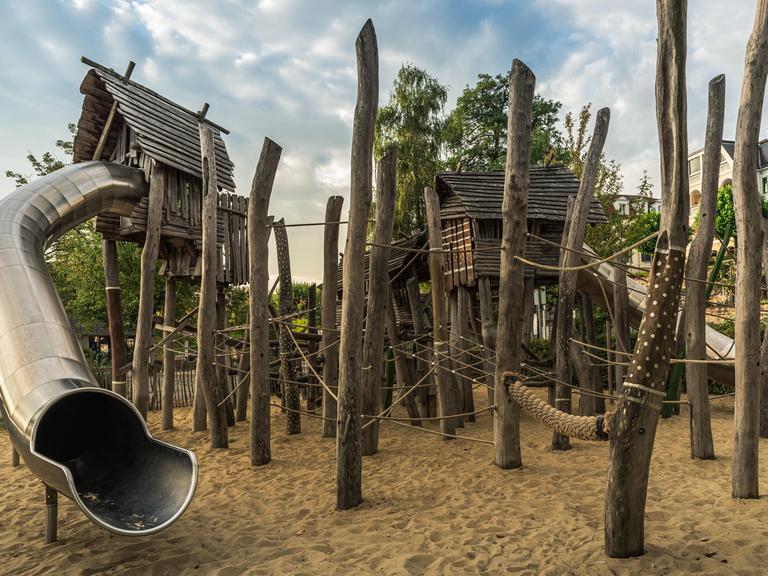 Ein Kinderspielplatz mit Holzhütten, Klettergeräten aus Holzpfählen und großzügiger Rutsche, der an einen Wald erinnert.  