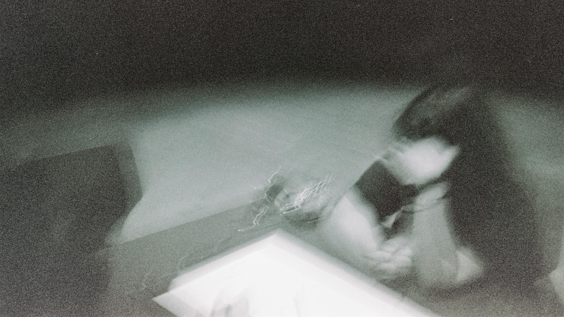 Ein verwackeltes Schwarz-Weiß-Bild von einer Frau, die an einem Tisch sitzt und sich eine Hand vors Gesicht hält.