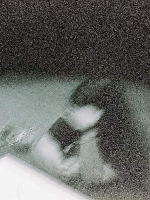 Ein verwackeltes Schwarz-Weiß-Bild von einer Frau, die an einem Tisch sitzt und sich eine Hand vors Gesicht hält.