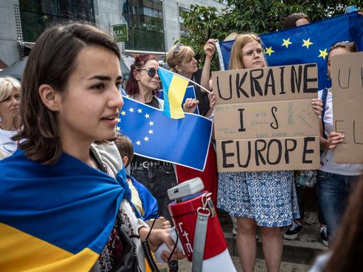 Teilnehmerinnen einer Protestaktion in Brüssel halten ukrainische Flaggen hoch und haben Plakate in der Hand